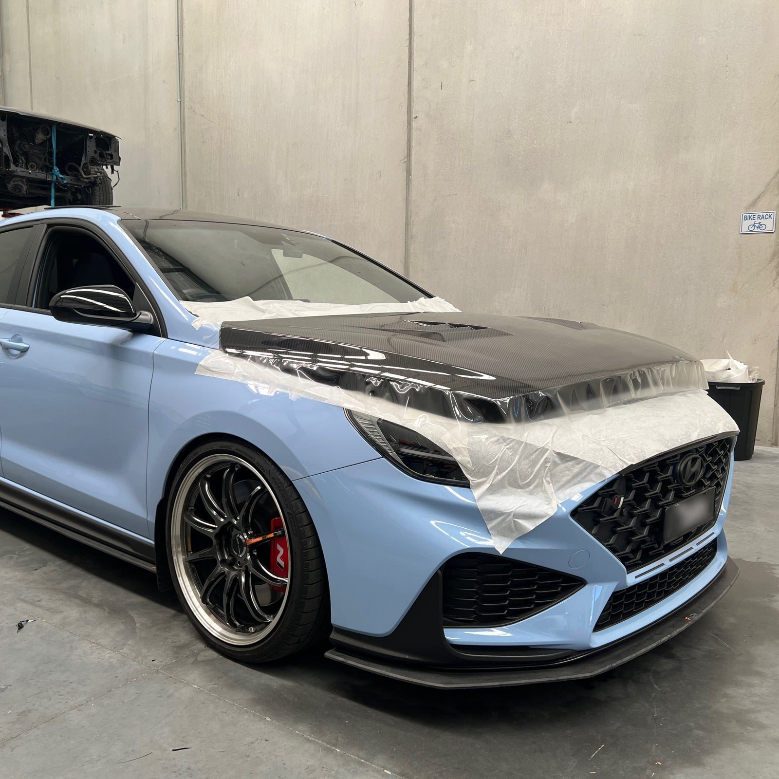 Paint protection film being laid over the carbon bonnet of a light blue Hyundai i30 N inside the Defiant Motor Culture workshop