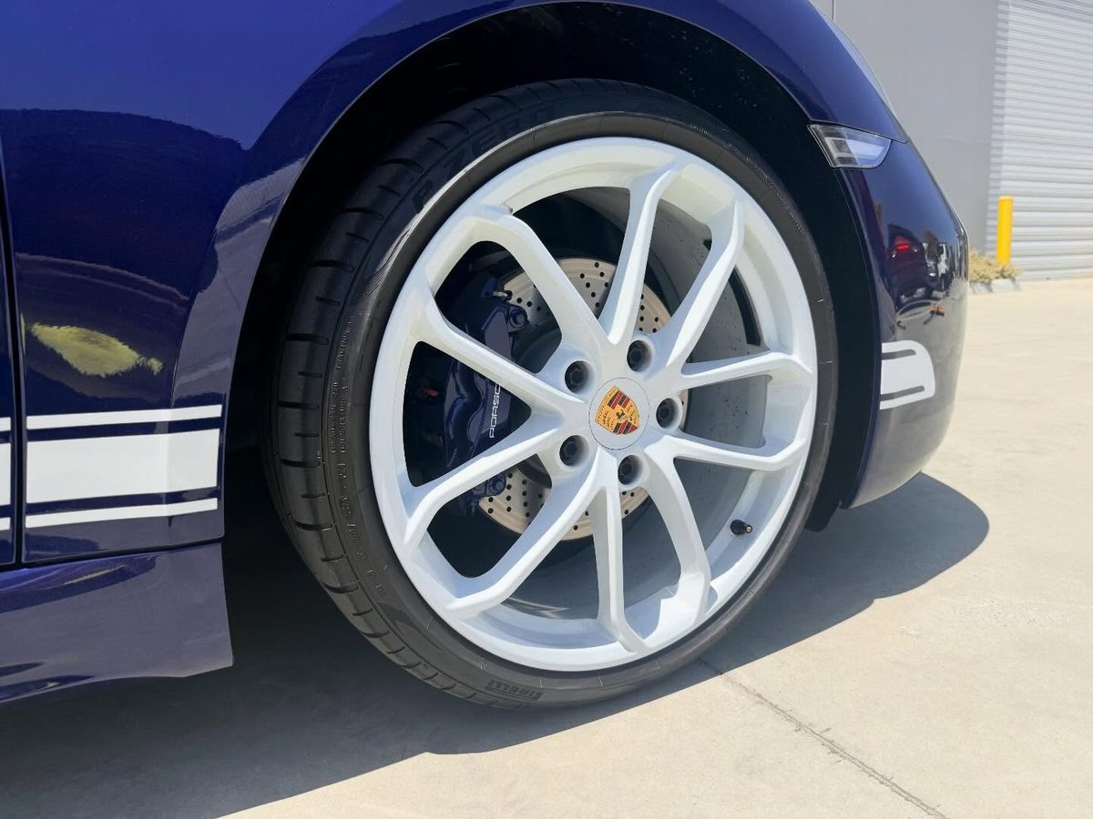 Close up of a Porsche 718 wheel finished in white, blue caliper behind it and white side stripes on the panel